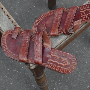 May include: A pair of brown leather sandals featuring a toe loop and two wide straps across the top. The footbed is decorated with a stamped geometric border pattern and a central embossed design, showcasing a textured, weathered finish throughout.