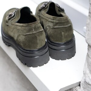 May include: A pair of olive green suede loafers featuring a thick black lug sole and a metallic horsebit detail across the top. The shoes have a rounded toe and a sturdy, chunky platform base, showcasing a textured suede finish and dark hardware.