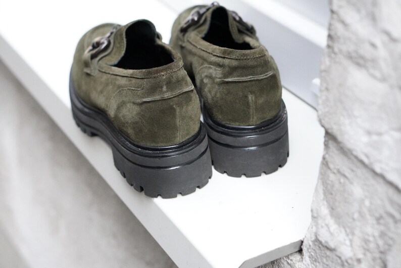 May include: A pair of olive green suede loafers featuring a thick black lug sole and a metallic horsebit detail across the top. The shoes have a rounded toe and a sturdy, chunky platform base, showcasing a textured suede finish and dark hardware.