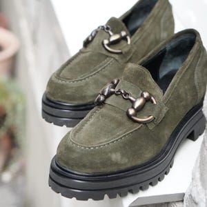 May include: A pair of olive green suede loafers featuring a chunky black lug sole and a metallic horsebit detail across the vamp. The shoes have a rounded toe design and a thick platform base, providing a sturdy and elevated look for casual footwear.