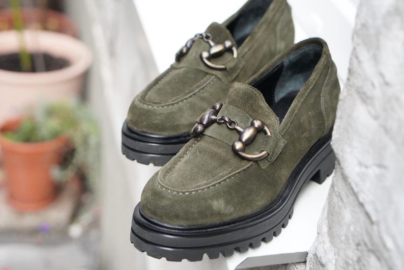 May include: A pair of olive green suede loafers featuring a chunky black lug sole and a metallic horsebit detail across the vamp. The shoes have a rounded toe design and a thick platform base, providing a sturdy and elevated look for casual footwear.
