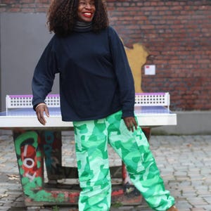 May include: A person wearing a navy blue long-sleeve shirt and green camouflage pants. The person is standing near a ping pong table with a white net and a colorful graffiti design on the base.