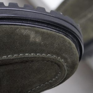May include: An olive green suede boot featuring a thick black rubber lug sole with deep treads. The upper part of the boot is crafted from soft olive green suede material with visible stitching along the toe cap and a metallic bronze hardware detail.