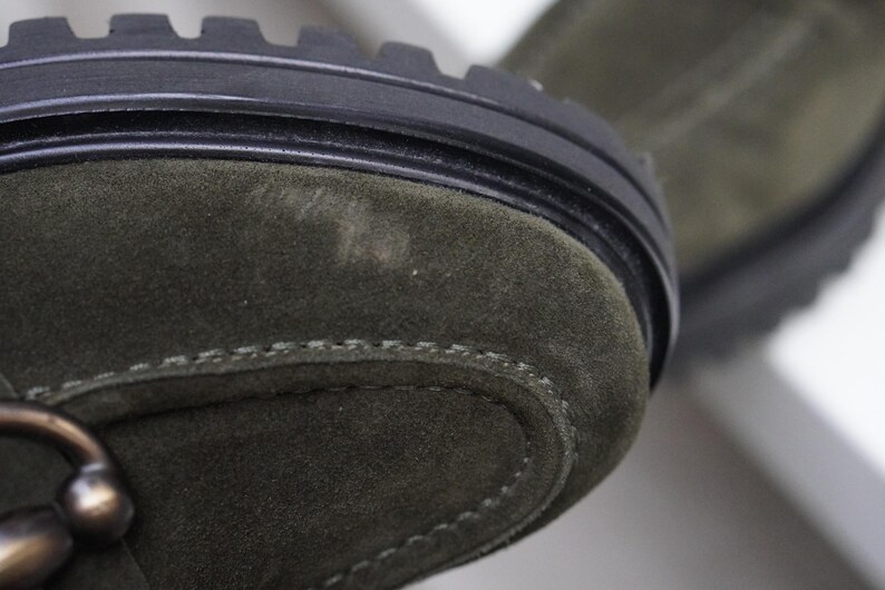 May include: An olive green suede boot featuring a thick black rubber lug sole with deep treads. The upper part of the boot is crafted from soft olive green suede material with visible stitching along the toe cap and a metallic bronze hardware detail.