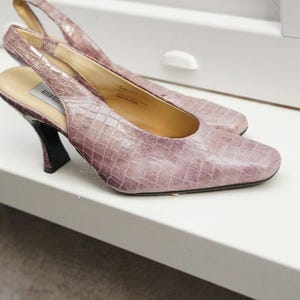 May include: A pair of mauve slingback heels with a square toe and a textured, reptile-like pattern. The heels have a black, curved heel and a strap that goes around the back of the ankle. The shoes are on a white surface.