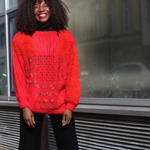 Könnte beinhalten: Ein leuchtend roter Pullover mit einem strukturierten Strickmuster und dekorativen Verzierungen. Der Pullover hat lange Ärmel und wird mit einer schwarzen Hose getragen. Ein schwarzer Rollkragen ist darunter sichtbar.