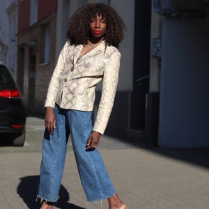 May include: A beige blazer with a snakeskin pattern, paired with blue denim jeans. The blazer has a classic cut with buttons. The jeans are wide-legged with a raw hem. The model is standing on a paved street.