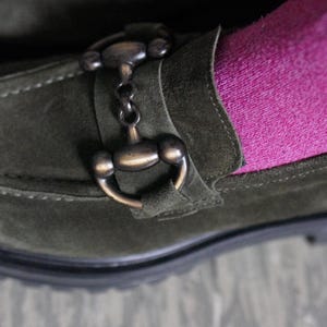 May include: An olive green suede loafer featuring a dark bronze-toned metal horsebit detail across the top. The shoe is worn with a bright pink sock, showcasing the contrast between the textured green suede and the vibrant fabric of the sock.