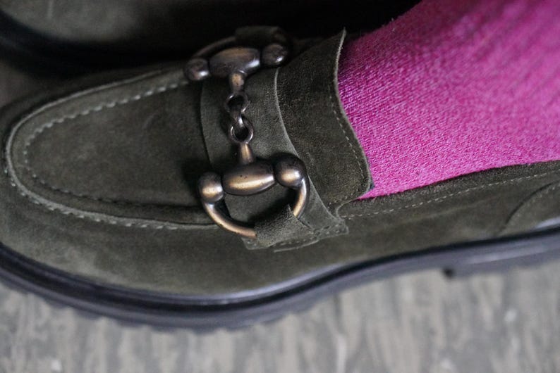May include: An olive green suede loafer featuring a dark bronze-toned metal horsebit detail across the top. The shoe is worn with a bright pink sock, showcasing the contrast between the textured green suede and the vibrant fabric of the sock.