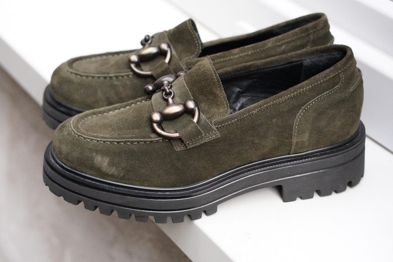 May include: A pair of olive green suede loafers featuring a dark metallic horsebit detail across the vamp. The shoes are designed with a thick, black, lugged rubber sole that provides a chunky silhouette and a prominent tread pattern for grip.