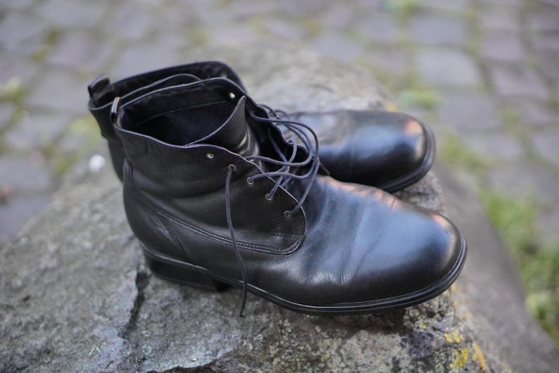 Op de afbeelding: Een paar zwarte leren laarzen met veters, rustend op een grijs stenen oppervlak. De laarzen hebben een ronde neus en een korte enkelhoogte. De veters zijn donker en het leer ziet er glad en gepolijst uit.