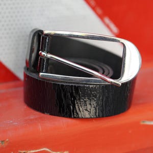 May include: A black leather belt with a silver-colored buckle. The belt is rolled up and sits on a bright orange surface. The buckle is rectangular with a rounded edge.