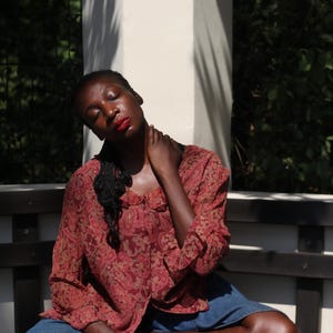 May include: A long-sleeved, button-down shirt in a reddish-brown hue featuring a repeating paisley pattern. The shirt has a rounded collar and is worn with blue denim bottoms. The fabric appears lightweight and semi-sheer with a loose, relaxed fit.