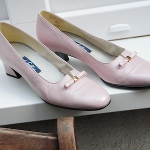 May include: A pair of light pink leather pumps with small heels and rounded toes. Each shoe features a small bow with a gold-colored accent. The shoes are displayed on a white surface.