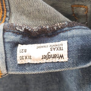 Pode incluir: Close-up de jeans azuis com uma etiqueta branca. A etiqueta diz "Wrangler TEXAS AUTHENTIC STRAIGHT" com o tamanho "31x30" e o estilo "821". Os jeans s&atilde;o feitos de tecido denim dur&aacute;vel.