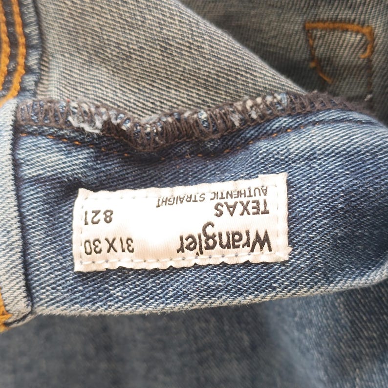 Pode incluir: Close-up de jeans azuis com uma etiqueta branca. A etiqueta diz "Wrangler TEXAS AUTHENTIC STRAIGHT" com o tamanho "31x30" e o estilo "821". Os jeans s&atilde;o feitos de tecido denim dur&aacute;vel.