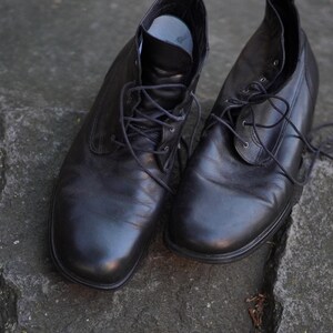 Op de afbeelding: Een paar zwarte leren enkellaarsjes met veters. De laarzen hebben een klassiek ontwerp, geschikt voor dagelijks gebruik. De schoenen staan op een getextureerd, grijs stenen oppervlak.