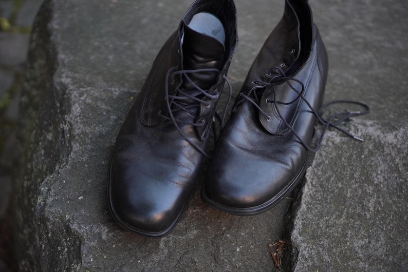 Op de afbeelding: Een paar zwarte leren enkellaarsjes met veters. De laarzen hebben een klassiek ontwerp, geschikt voor dagelijks gebruik. De schoenen staan op een getextureerd, grijs stenen oppervlak.