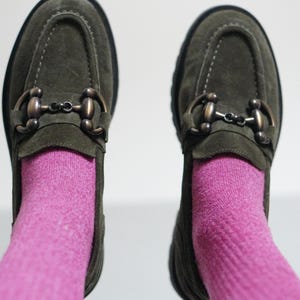 May include: A pair of olive green suede loafers featuring a dark bronze-toned metal horsebit detail across the top. The shoes are worn with bright pink socks, showcasing a moccasin-style toe construction and a thick, dark-coloured rubber sole.