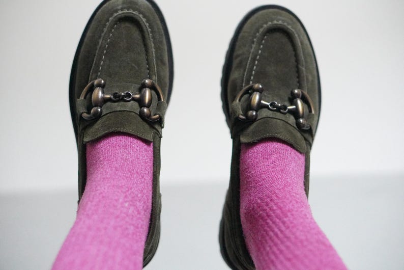 May include: A pair of olive green suede loafers featuring a dark bronze-toned metal horsebit detail across the top. The shoes are worn with bright pink socks, showcasing a moccasin-style toe construction and a thick, dark-coloured rubber sole.