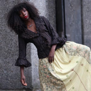 Op de afbeelding: Een zwarte blouse met ruchesmouwen en een diepe halslijn wordt gecombineerd met een lange, zwierige rok. De rok is lichtgeel met een bloemenpatroon en geborduurde details. De outfit wordt buiten gedragen.