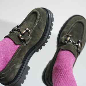 May include: A pair of olive green suede loafers featuring a chunky black lug sole and a metallic horsebit detail across the top. The shoes are worn with bright pink ribbed socks, showcasing the textured suede material and the thick, treaded rubber outsole.