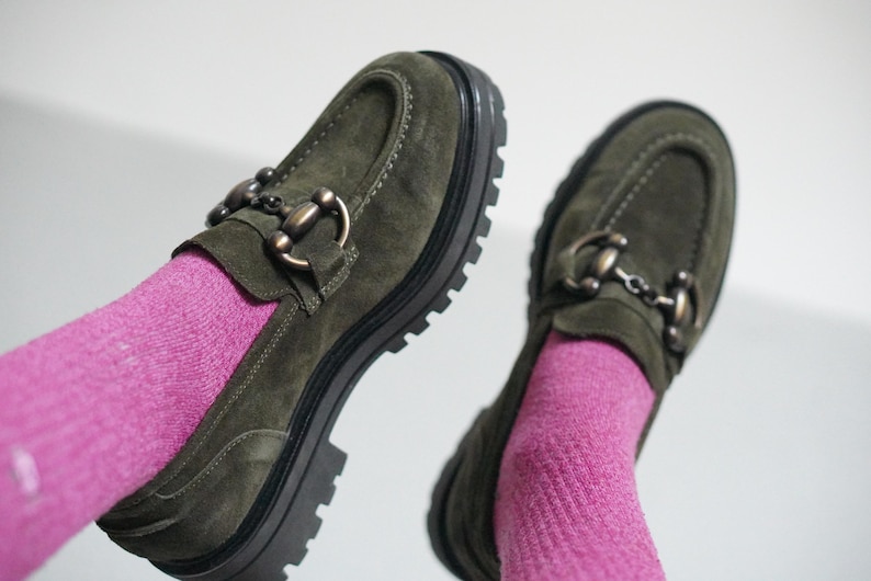 May include: A pair of olive green suede loafers featuring a chunky black lug sole and a metallic horsebit detail across the top. The shoes are worn with bright pink ribbed socks, showcasing the textured suede material and the thick, treaded rubber outsole.