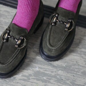 May include: A pair of olive green suede loafers featuring a prominent bronze-toned metal horsebit detail across the vamp. The shoes have a thick black rubber lug sole and are worn with bright pink socks that extend above the ankle line.