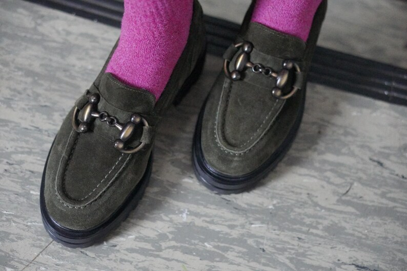 May include: A pair of olive green suede loafers featuring a prominent bronze-toned metal horsebit detail across the vamp. The shoes have a thick black rubber lug sole and are worn with bright pink socks that extend above the ankle line.