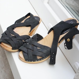 Może przedstawiać: Para czarnych skórzanych chodaków z plecionym wzorem z przodu. Chodaki mają drewnianą podeszwę i mały obcas. Pasek na kostkę jest regulowany. Buty są prezentowane na białej powierzchni.