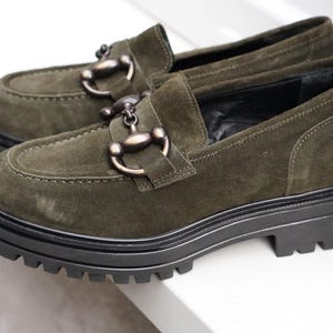 May include: A pair of olive green suede loafers featuring a dark metallic horsebit detail across the vamp. The shoes are designed with a thick, black, lugged rubber sole that provides a chunky silhouette and a prominent tread pattern for grip.
