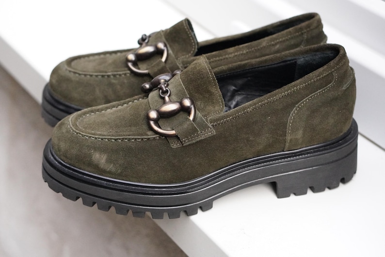May include: A pair of olive green suede loafers featuring a dark metallic horsebit detail across the vamp. The shoes are designed with a thick, black, lugged rubber sole that provides a chunky silhouette and a prominent tread pattern for grip.