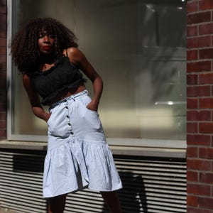 Könnte beinhalten: Ein schwarzes Crop-Top mit Rüschensaum und ein hellblauer Jeansrock mit großen schwarzen Knöpfen und Taschen. Der Rock hat einen Rüschensaum. Das Model steht vor einer Backsteinmauer und einem Fenster.