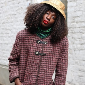 Könnte beinhalten: Eine Person trägt einen Strohhut und eine gemusterte Jacke mit schwarzen Lederverschlüssen. Die Jacke ist eine Mischung aus Braun- und Rottönen. Ein grün-schwarz gestreifter Rollkragenpullover ist sichtbar.