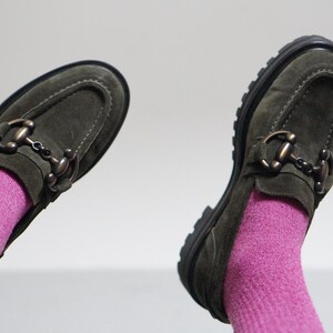 May include: A pair of olive green suede loafers featuring a metallic horsebit detail across the vamp, visible stitching along the toe, and a thick black lug sole. The shoes are worn with bright pink ribbed socks against a plain light background.
