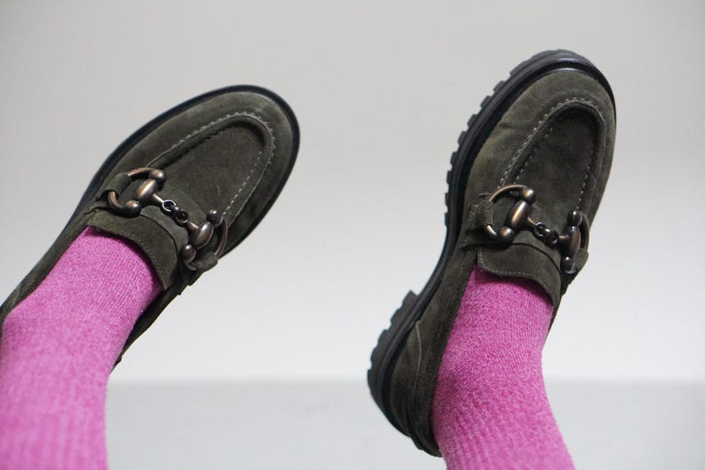 May include: A pair of olive green suede loafers featuring a metallic horsebit detail across the vamp, visible stitching along the toe, and a thick black lug sole. The shoes are worn with bright pink ribbed socks against a plain light background.