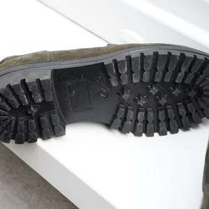 May include: An olive green suede loafer with a thick black rubber lug sole featuring a deep tread pattern. The shoe is positioned to show the underside of the sole, which has a block heel and a textured grip design for traction on various surfaces.