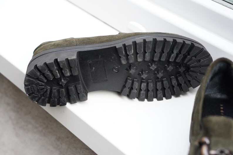May include: An olive green suede loafer with a thick black rubber lug sole featuring a deep tread pattern. The shoe is positioned to show the underside of the sole, which has a block heel and a textured grip design for traction on various surfaces.