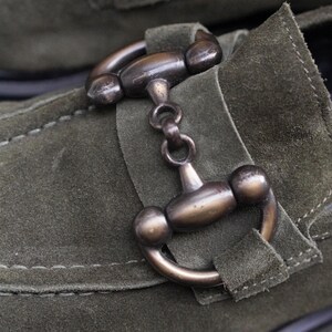 May include: An olive green suede loafer featuring a prominent metallic horsebit detail in a dark bronze finish. The shoe is constructed with visible tonal stitching along the vamp and a structured strap that secures the metal hardware in place.
