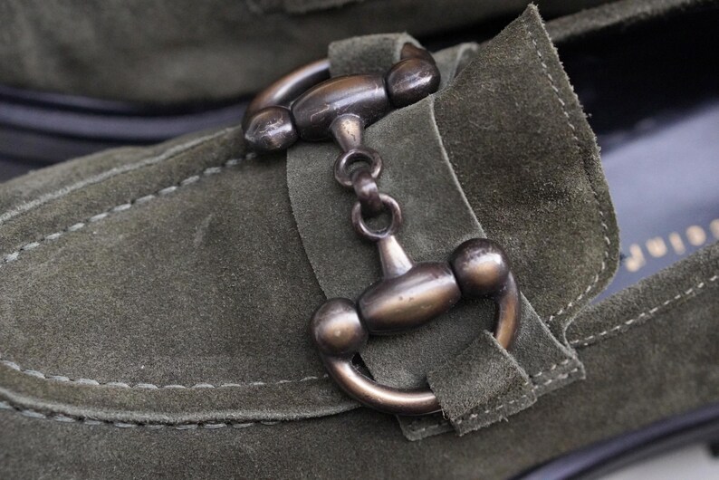 May include: An olive green suede loafer featuring a prominent metallic horsebit detail in a dark bronze finish. The shoe is constructed with visible tonal stitching along the vamp and a structured strap that secures the metal hardware in place.