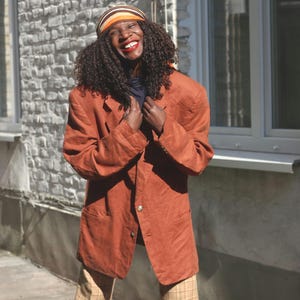 Könnte beinhalten: Ein rostfarbener Blazer mit einem einzelnen Knopfverschluss. Der Blazer wird über einem dunkelblauen Oberteil und mit einer karierten Hose getragen. Ein gestreiftes Stirnband in Orange- und Brauntönen wird auf dem Kopf getragen.