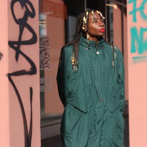 Può includere: Una persona indossa un cappotto verde scuro a maniche lunghe con chiusura a bottoni. La persona ha lunghi capelli intrecciati con perline decorative. Lo sfondo presenta graffiti su un muro rosa.