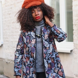Op de afbeelding: Een vrouw draagt een jas met een bloemenpatroon in blauw, rood en paars. De jas is open en onthult een grijze coltrui. Ze draagt ook een bruine hoed en een ketting.