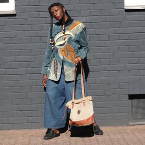 Könnte beinhalten: Eine blaugrüne Wildlederjacke mit großen beigen und cremefarbenen blattförmigen Applikationen, kombiniert mit einer weiten blauen Jeanshose und einer beigen Canvas-Tasche mit braunem Lederbesatz, einem schwarzen Rollkragenpullover und schwarzen Schuhen.