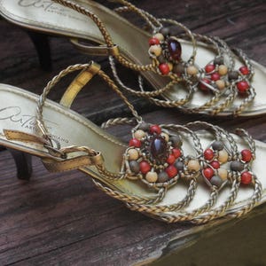 May include: A pair of gold-toned sandals with a kitten heel, featuring braided straps across the foot. The design is embellished with a central cluster of round red, brown, and cream-colored beads, with a dark brown oval stone at the center.