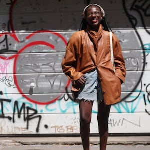 May include: A tan leather jacket with a high collar and button-down front, paired with light blue denim shorts featuring a raw, frayed hemline. The jacket has long sleeves and a loose, relaxed fit, worn over the denim shorts.