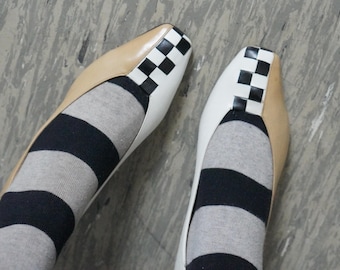 May include: A pair of square-toe flats featuring a colour-blocked design with tan leather sides, white leather centres, and a black and white checkered pattern on the toe caps. The shoes are worn with grey and black horizontal striped socks.