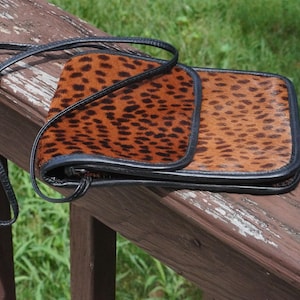 Könnte beinhalten: Eine rechteckige Umhängetasche mit braun-schwarzem Leopardenmuster auf einer texturierten Oberfläche in Felloptik. Die Tasche verfügt über einen dünnen schwarzen Lederriemen und schwarze Lederpaspeln an den Kanten der Klappe und des Hauptteils.