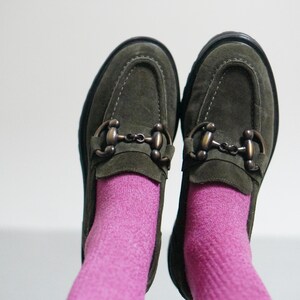 May include: A pair of olive green suede loafers featuring a dark bronze-toned metal horsebit detail across the top. The shoes are worn with bright pink socks, showcasing a rounded toe design and a thick black sole with visible stitching.