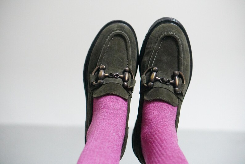 May include: A pair of olive green suede loafers featuring a dark bronze-toned metal horsebit detail across the top. The shoes are worn with bright pink socks, showcasing a rounded toe design and a thick black sole with visible stitching.